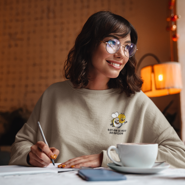 Young professional wearing the oatmeal cream crewneck sweatshirt while sitting in a cafe with a coffee cup, highlighting the "9:01 AM IS NOT AN OPTION" bee graphic.