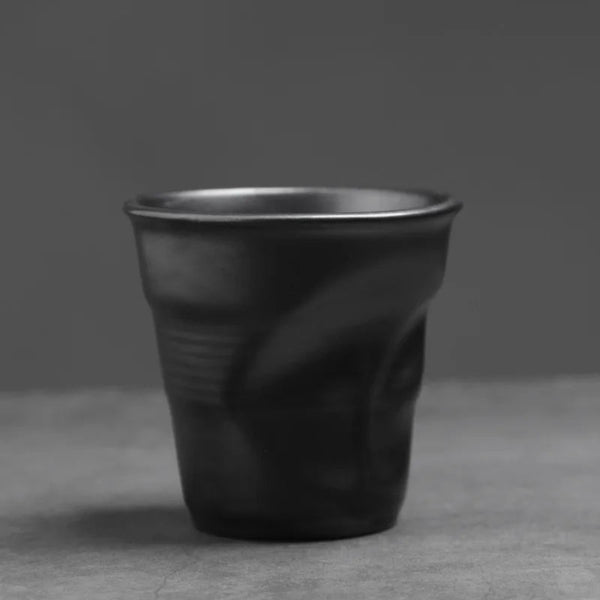 Matte black crumpled ceramic espresso cup with a hand-pinched pleated texture sitting on a grey surface.