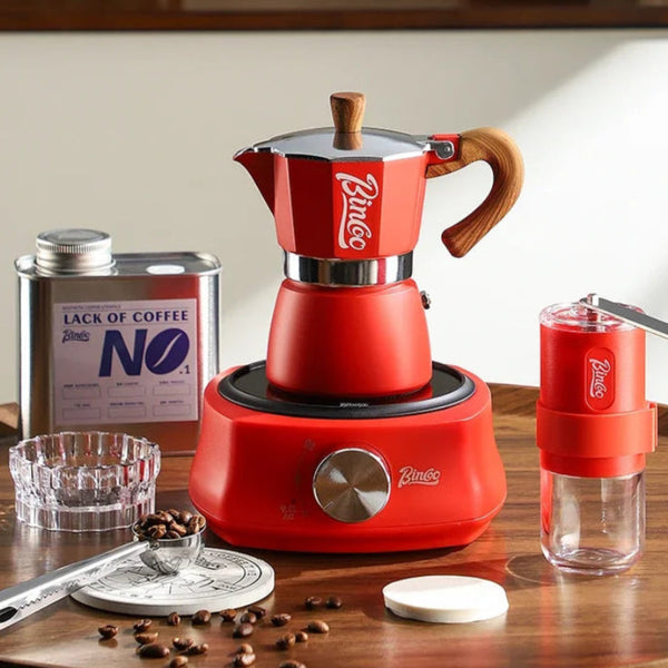 A red Bincoo moka pot coffee maker set on a wooden table, shown with a matching red manual grinder, a metal coffee tin labeled 'Lack of Coffee No. 1', coffee beans, a glass ashtray, and a small scale.