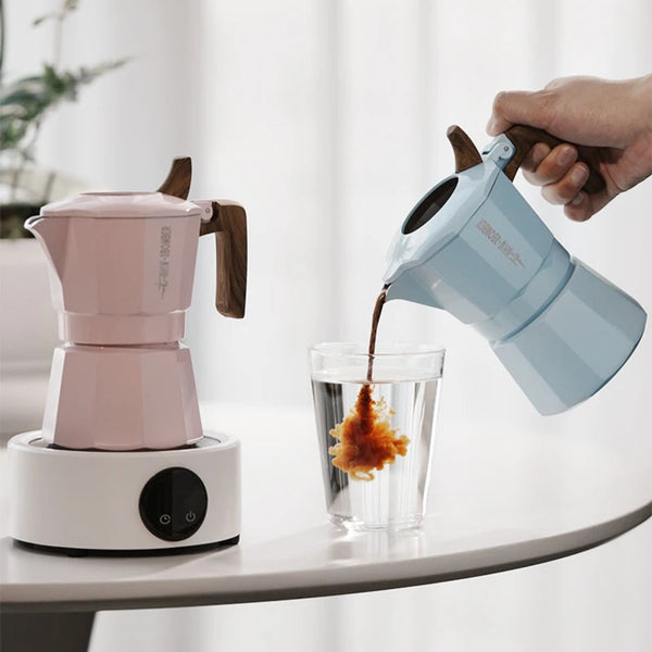 A light blue and white dual valve moka pot is shown being filled with dark coffee and poured into a glass on a white table, with a pink electric burner beside it. Text at the top reads 'DUAL VALVE EXTRACTION RICH CREMA' and 'Flipped Moka Pot'.