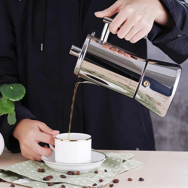 Person pouring coffee from a stainless steel Moka Pot into a white mug on a table with scattered coffee beans.
