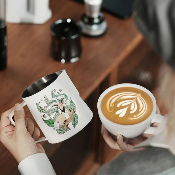 MHW-3BOMBER Origin Series milk pitcher held next to a finished cup of professional latte art on a wooden coffee bar.