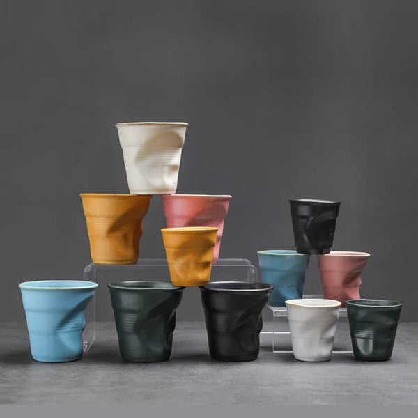 A group display of handmade crumpled ceramic coffee cups in black, blue, pink, white, and yellow against a grey background.