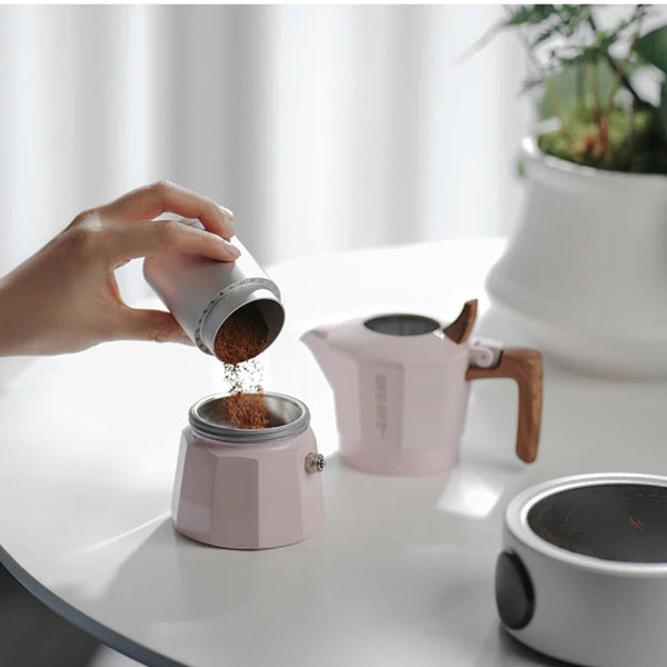 A hand pours coffee grounds from a small white and silver pour-over coffee maker into a pink moka pot on a white table. A matching pink kettle and a white electric burner sit in the background near a potted plant.