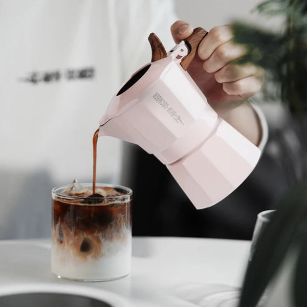 A light pink moka pot with a dark wood handle is shown pouring coffee into an iced latte in a glass cup, with a person's hand holding the pot in a kitchen setting.