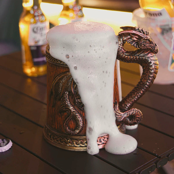 Cold beer overflowing with thick white foam from a classic bronze medieval dragon barrel tankard on a dark wooden patio table.