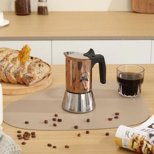 Rose gold and stainless steel moka pot stovetop espresso maker on kitchen counter next to a glass of coffee and sliced bread.