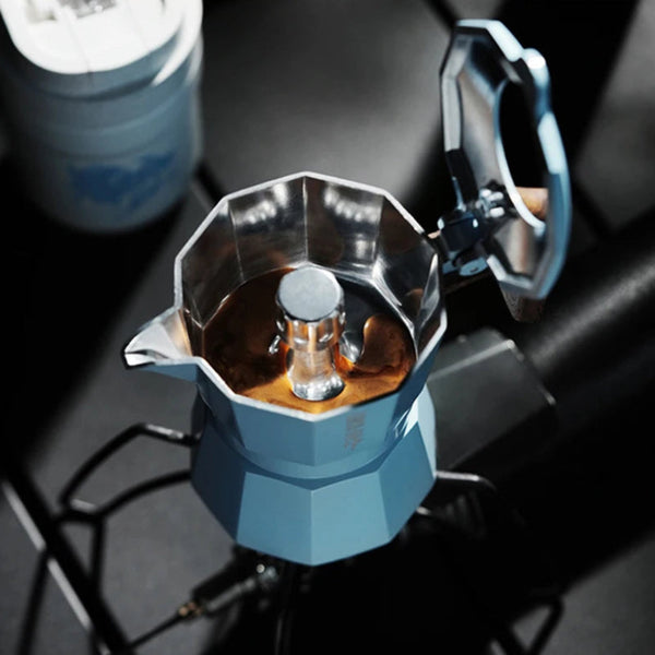 A blue and stainless steel moka pot espresso maker on a black burner, with coffee visible inside and its lid open.