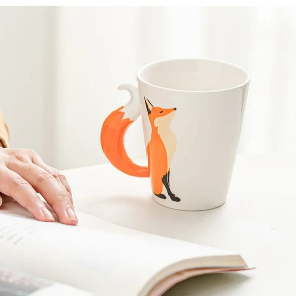 Cozy lifestyle view of the standing fox ceramic mug resting on a white table next to a person reading an open book.