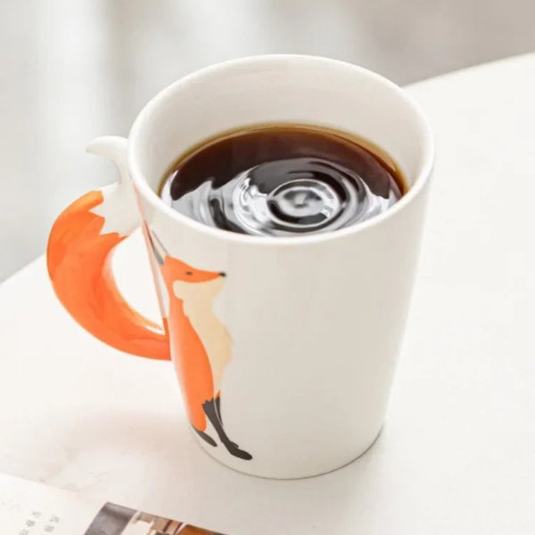 Top-down view of a white ceramic fox mug filled with hot black coffee, highlighting the orange fox illustration and 3D tail handle on a bright surface.
