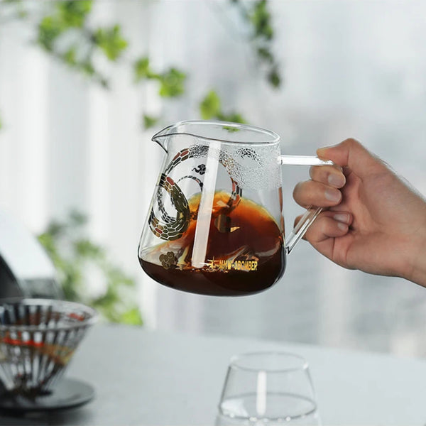 Hand holding an MHW-3BOMBER Serpent Series 500ml Glass Coffee Server filled with brewed coffee in a natural light setting.