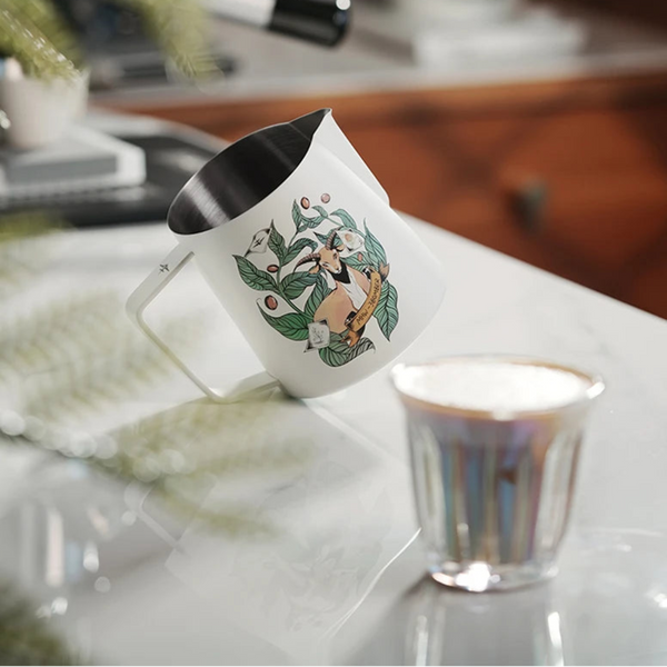MHW-3BOMBER Origin Series white milk pitcher on a marble countertop next to a glass of freshly steamed milk.