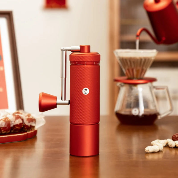 TIMEMORE Chestnut S3 Red manual coffee grinder standing upright on a wooden table, featuring a matching red gooseneck kettle pouring water into a glass pour-over coffee dripper set up in the blurred background.