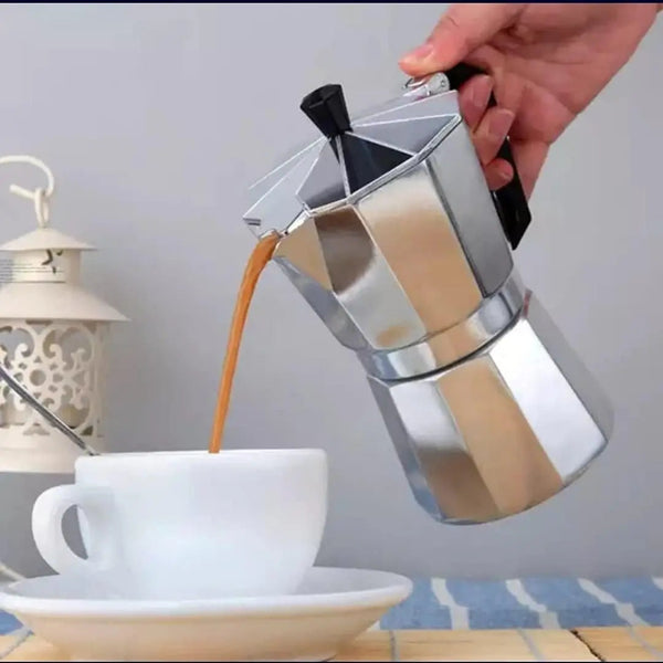 A hand holds a stainless steel moka pot and pours espresso into a white ceramic cup on a saucer, with a decorative lantern in the background.