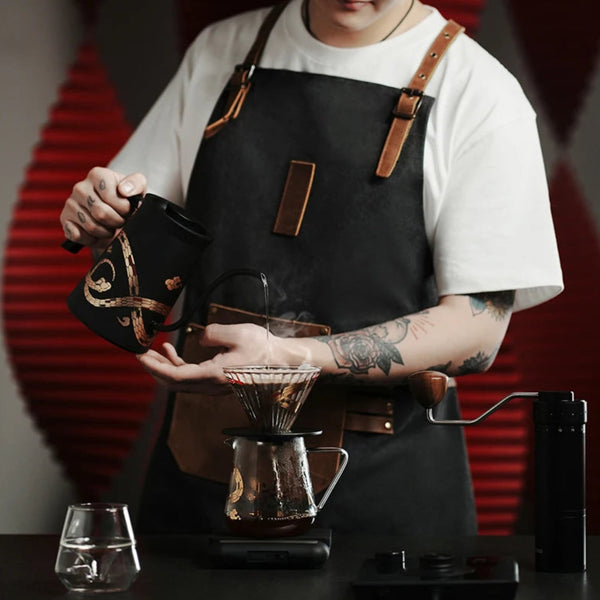 Professional barista using the MHW-3BOMBER Serpent Series pour-over kit, featuring the black gooseneck kettle, glass coffee server, and manual hand grinder.