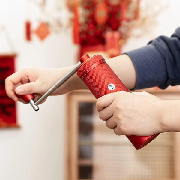 Action shot of a user operating the TIMEMORE Chestnut S3 Red manual coffee grinder, turning the extended silver metal crank arm with one hand while firmly gripping the textured red aluminum unibody with the other.