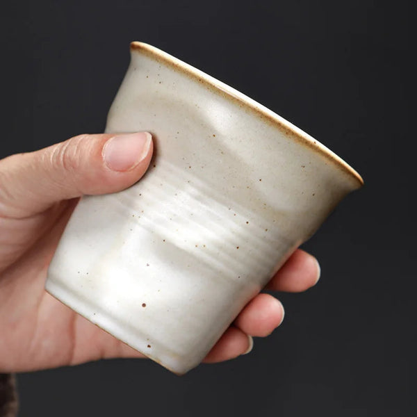 A hand holding a white crumpled ceramic espresso cup to show the ergonomic pleated texture and physical scale against a dark background.