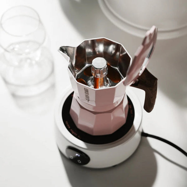A pink stovetop moka pot with its lid open, sitting on a white electric burner. The brand name 'MOCAS' is visible on the side of the pot.