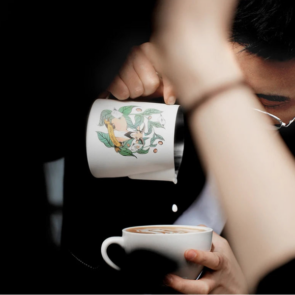 Close-up of a barista using an MHW-3BOMBER Origin Series pitcher to pour milk and create detailed latte art.


