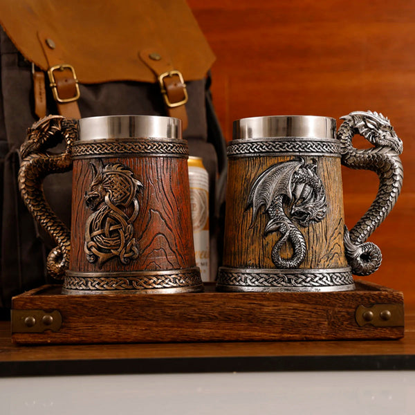 Front-facing view of the complete medieval dragon barrel tankard collection, displaying the dark bronze variant with its roaring dragon crest next to the antique silver variant with its coiled dragon wings on a wooden tray.