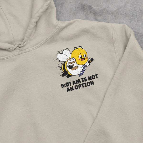 A close-up view of an Oatmeal Cream beige unisex hooded sweatshirt, highlighting a detailed left-chest embroidery placement. The design features a running corporate bee mascot dressed in a collared work shirt and necktie, frantically gripping a coffee cup and checking his watch, with the stacked text slogan 9:01 AM IS NOT AN OPTION stitched cleanly underneath.