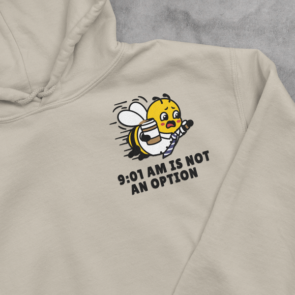 A close-up view of an Oatmeal Cream beige unisex hooded sweatshirt, highlighting a detailed left-chest embroidery placement. The design features a running corporate bee mascot dressed in a collared work shirt and necktie, frantically gripping a coffee cup and checking his watch, with the stacked text slogan 9:01 AM IS NOT AN OPTION stitched cleanly underneath.
