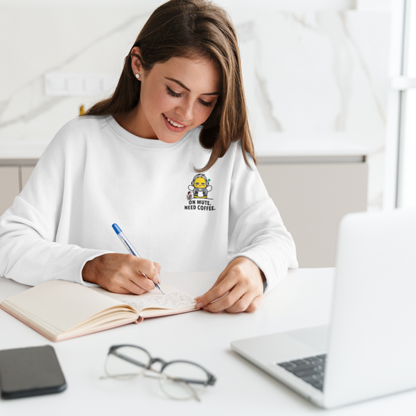 A smiling woman sitting at a desk with a laptop, writing in a notebook while wearing a ceramic white sweatshirt featuring a funny "On Mute. Need Coffee." corporate bee design.
