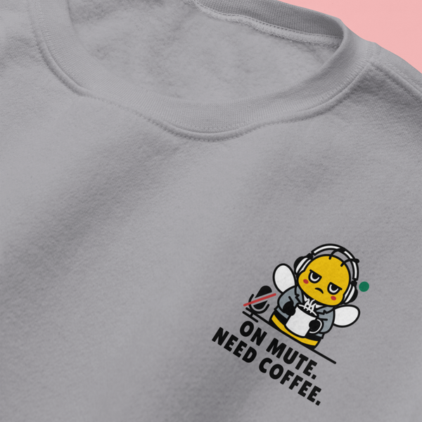 Close-up flat lay of a heather ash grey crewneck sweatshirt on a pink background, showcasing the embroidered details of a funny corporate bee mascot reading "On Mute. Need Coffee."