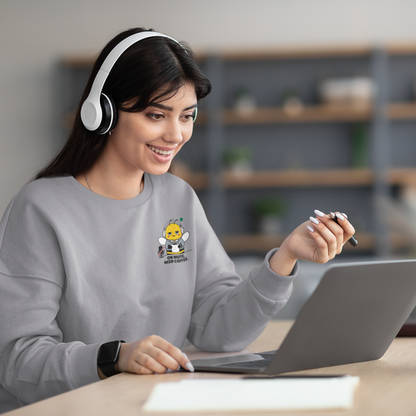 A smiling woman wearing white headphones and a heather ash grey crewneck sweatshirt with a funny "On Mute. Need Coffee." bee design, looking at her laptop during a virtual video call.