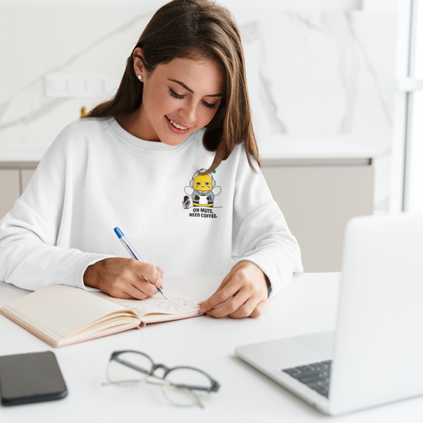 A smiling woman sitting at a desk with a laptop, writing in a notebook while wearing a ceramic white sweatshirt featuring a funny "On Mute. Need Coffee." corporate bee design.