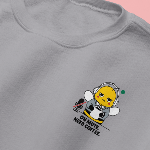 Close-up flat lay of a heather ash grey crewneck sweatshirt on a pink background, showcasing the embroidered details of a funny corporate bee mascot reading "On Mute. Need Coffee."