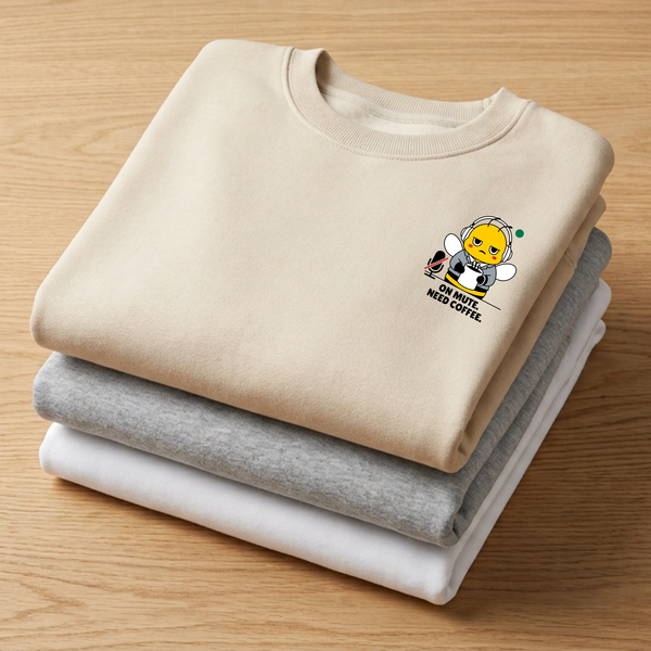 •  Stack of folded crewneck sweatshirts in oatmeal cream, heather ash grey, and ceramic white on a wooden table, showcasing a beige sweatshirt with a funny mascot bee graphic reading "On Mute. Need Coffee."
