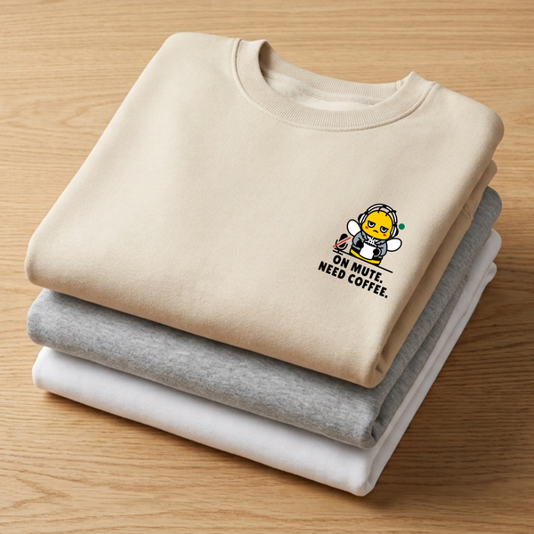 Stacks of folded crewneck sweatshirts in oatmeal cream, heather ash grey, and ceramic white on a wooden table, showcasing a beige sweatshirt with a funny mascot bee graphic reading "On Mute. Need Coffee."
