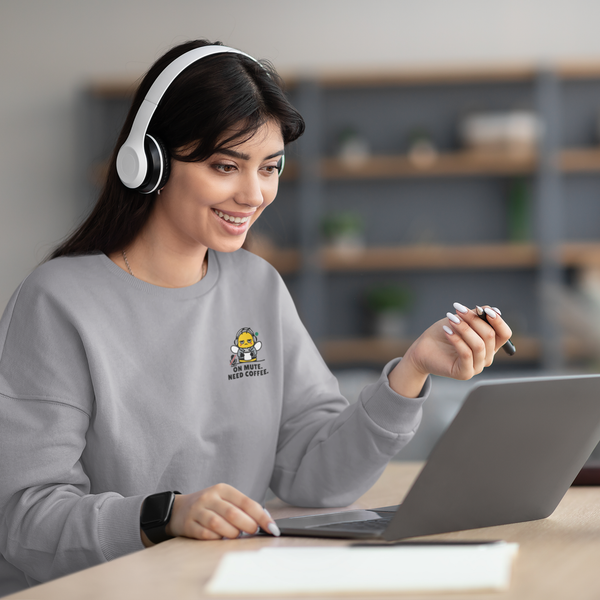 A smiling woman wearing white headphones and a heather ash grey crewneck sweatshirt with a funny "On Mute. Need Coffee." bee design, looking at her laptop during a virtual video call.
