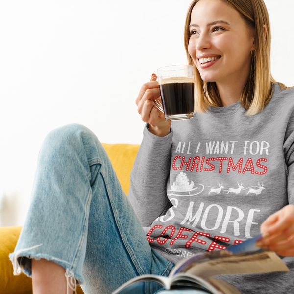 A woman wearing a sport grey Gildan 18000 Christmas sweatshirt that reads "All I want for Christmas is more coffee, Starbrew" sitting on a safa sipping coffee.