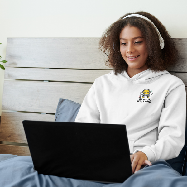 A smiling young woman sitting in bed with white headphones, working on a black laptop while wearing a ceramic white pullover hoodie featuring a small left-chest graphic of a cartoon bee and the text "ON MUTE. NEED COFFEE."