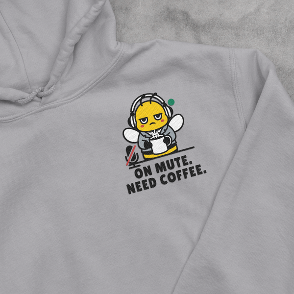 A close-up view of an Oatmeal Cream beige unisex hooded sweatshirt, highlighting a detailed left-chest embroidery placement. The design features a running corporate bee mascot dressed in a collared work shirt and necktie, frantically gripping a coffee cup and checking his watch, with the stacked text slogan ON MUTE. NEED COFFEE.  stitched cleanly underneath.