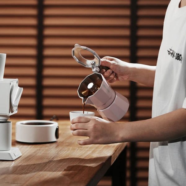 A person pouring coffee from a white stovetop moka pot into a small cup on a wooden table, with a coffee maker and another cup in the background against a brown slatted wall.