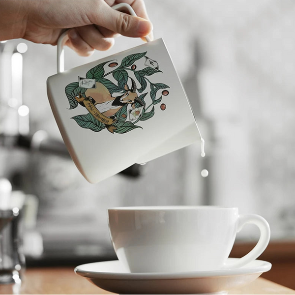 Hand pouring milk from an MHW-3BOMBER Origin Series pitcher into a white espresso cup for latte art.