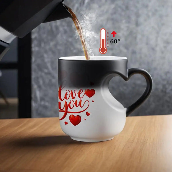 A heart-shaped ceramic mug with a black-to-white gradient, featuring red 'love you' script and heart graphics. A coffee pot is pouring hot coffee into the mug, with a digital temperature overlay indicating 60 degrees Celsius.