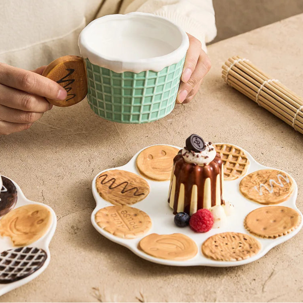 Mint green waffle texture ceramic mug with cookie handle held by hands, paired with a flower-shaped dessert plate featuring miniature ceramic biscuit decorations.