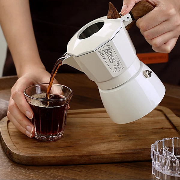 Person pouring coffee from a white coffee maker into a glass on a wooden table, with 'Binco' branding visible.
