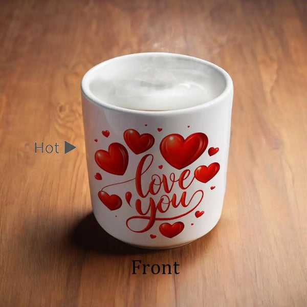 A white ceramic mug filled with a white beverage sits on a wooden tabletop. The mug is decorated with red hearts and the phrase 'love you' in red script.