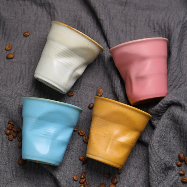 Overhead view of four crumpled ceramic coffee cups in white, pink, blue, and yellow scattered with roasted coffee beans on a textured grey fabric.