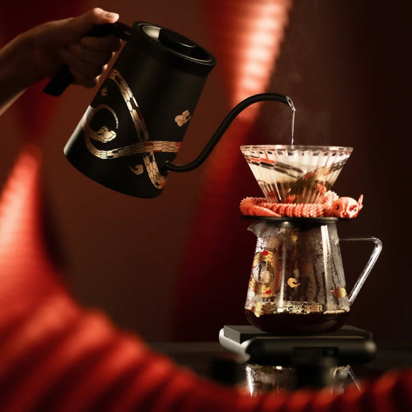 A barista in traditional attire preparing pour-over coffee with the MHW-3BOMBER Serpent Series collection in a festive red setting.