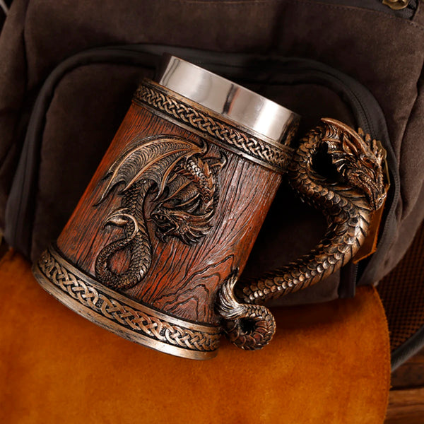 Overhead angled view of the classic bronze medieval dragon barrel tankard resting on a leather and canvas vintage bag, showcasing the detailed metallic dragon tail handle and braided trim.