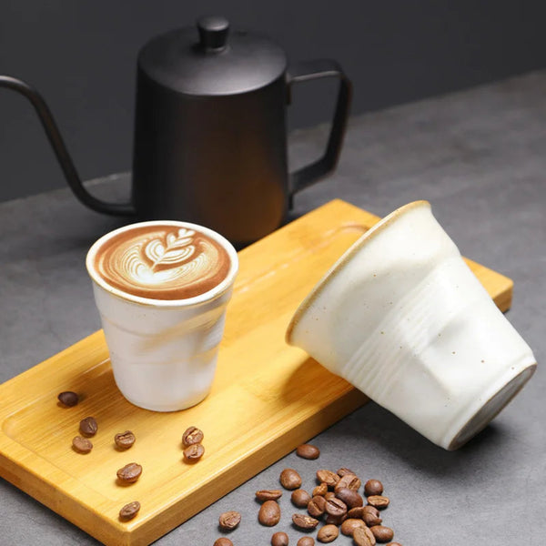 White hand-pinched pleated stoneware cup filled with fresh cappuccino latte art sitting on a wooden serving tray next to a black pour-over kettle.