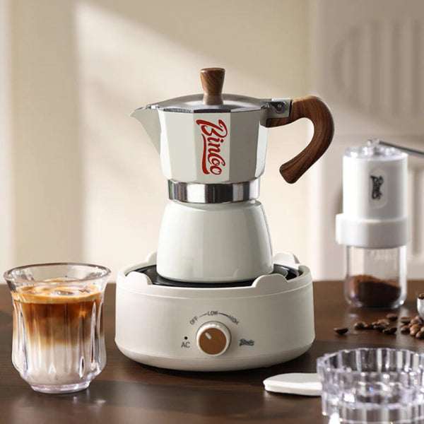 A white and silver moka pot with a wooden handle and lid knob sits on a matching white electric coffee maker base. A glass of iced coffee is on the left, and a coffee grinder and coffee beans are on the right, all set on a wooden table in a warm indoor setting.