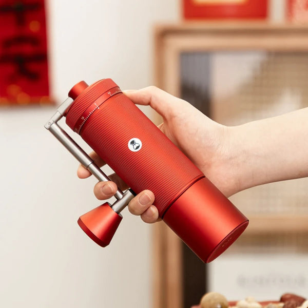 A person's hand holding the TIMEMORE Chestnut S3 Red manual coffee grinder at an angle, demonstrating its ergonomic grip size, textured aluminum unibody, and patented metal foldable handle mechanism in a warm indoor home environment.