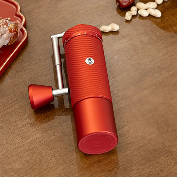 TIMEMORE Chestnut S3 Crimson Red manual coffee grinder resting flat on a polished wooden table, styled in a lifestyle home setup with a decorative red tray and scattered nuts alongside the textured red aluminum body and foldable metal handle.